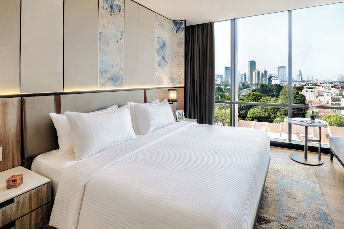 Une chambre d’hôtel avec un grand lit, une table de chevet, une petite table et une vue sur le paysage urbain grâce à de grandes fenêtres.