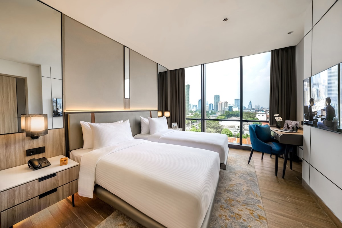 Une chambre d’hôtel avec deux lits, un bureau, une chaise, une grande fenêtre donnant sur la ville et un miroir.