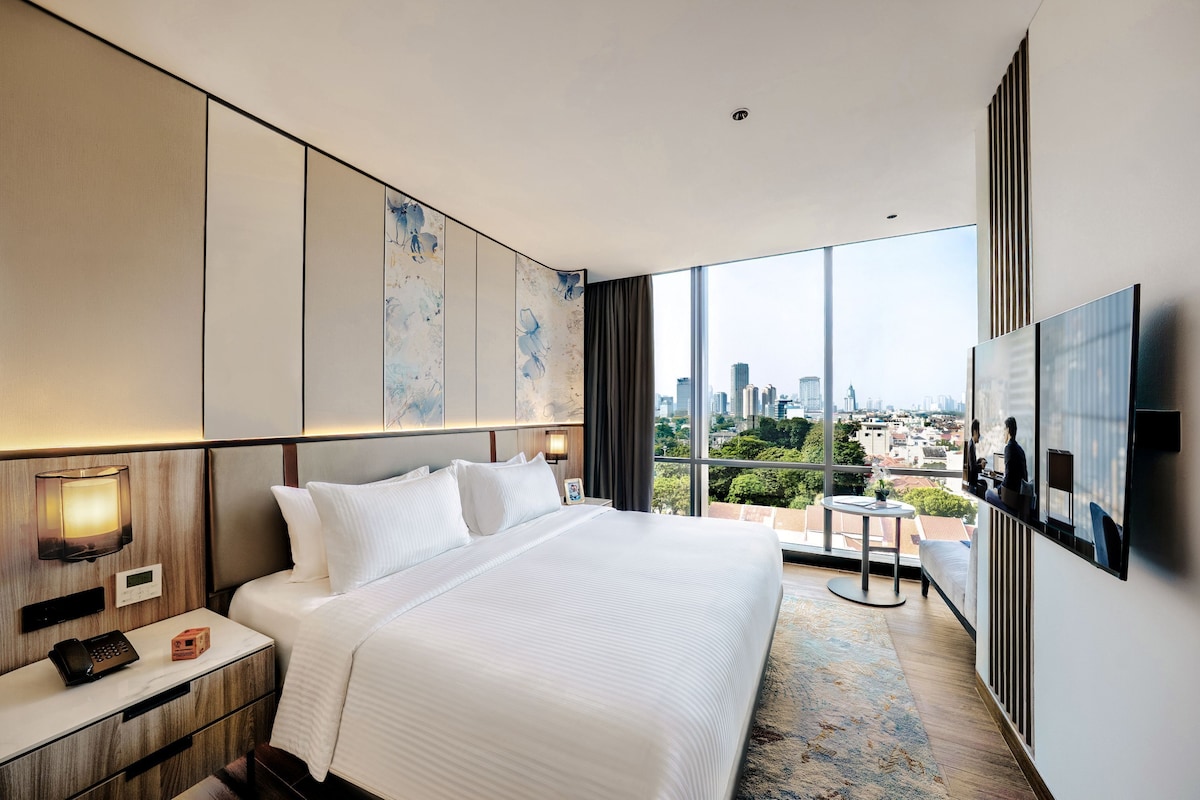 Une chambre d’hôtel moderne dotée d’un grand lit, d’une table de chevet avec un téléphone, d’un téléviseur à écran plat et offrant une vue sur la ville grâce à de grandes fenêtres.