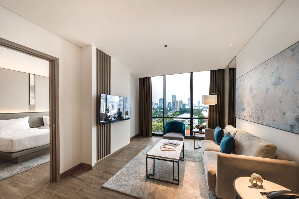 Une chambre d’hôtel moderne équipée d’un lit, d’un canapé, d’une table basse, d’une télévision et d’une grande fenêtre donnant sur la ville.