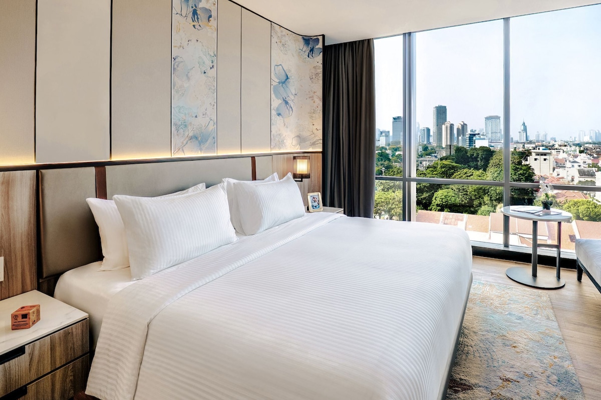 Une chambre d’hôtel avec un grand lit, une table de chevet, une petite table et une vue sur le paysage urbain grâce à de grandes fenêtres.
