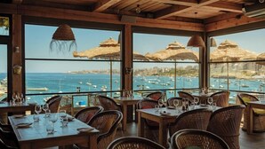 A restaurant with wooden furniture, large windows, and thatched umbrellas overlooking a marina. - Ushuaia Playa Xemxija Boutique Hotel (St. Paul's Bay)