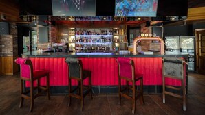 Zona de bar con mostrador rojo, varias estanterías llenas de botellas y tres taburetes.