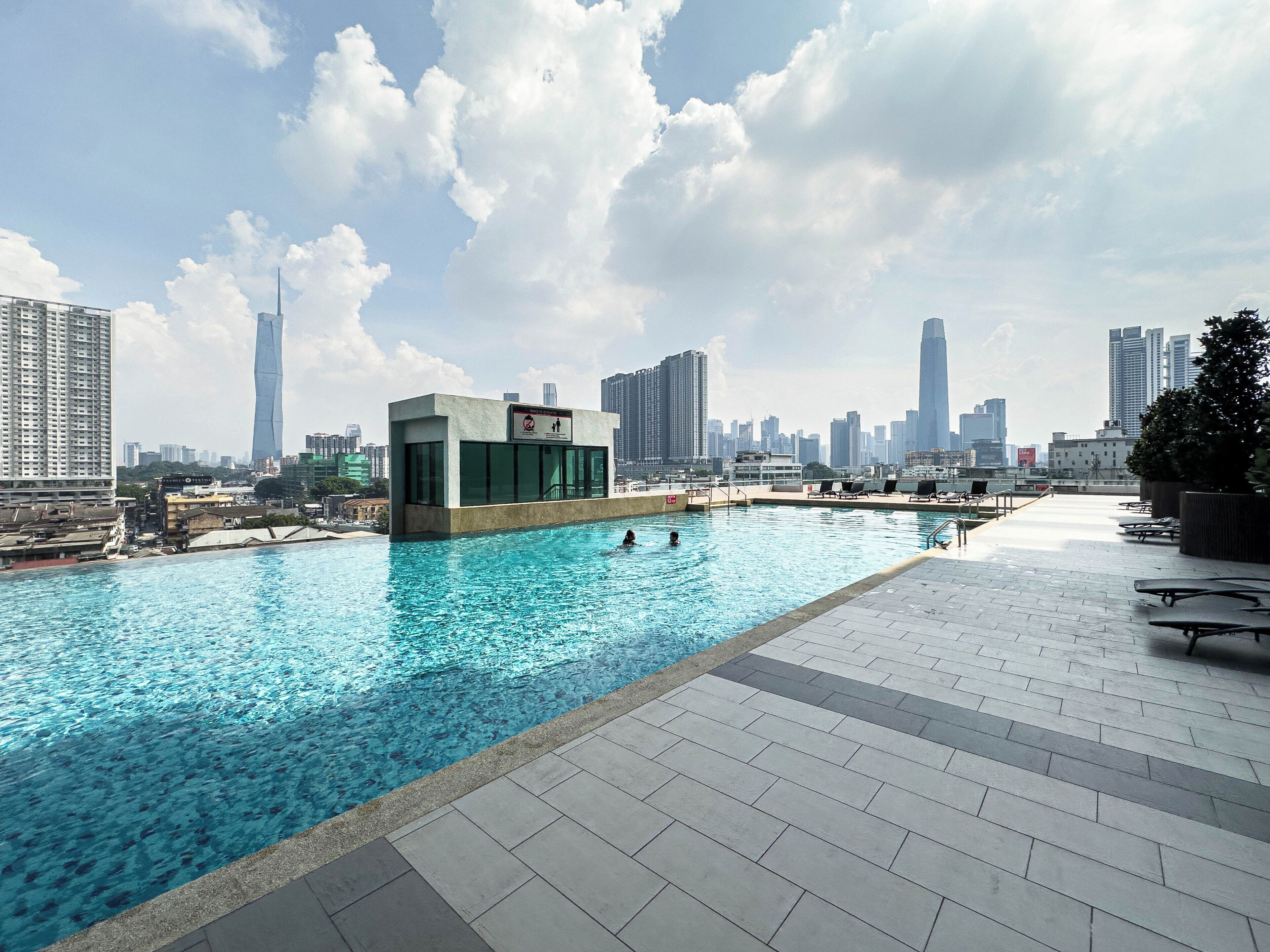 Una piscina en la azotea con un horizonte urbano de fondo, un edificio pequeño con un letrero y un cielo despejado.