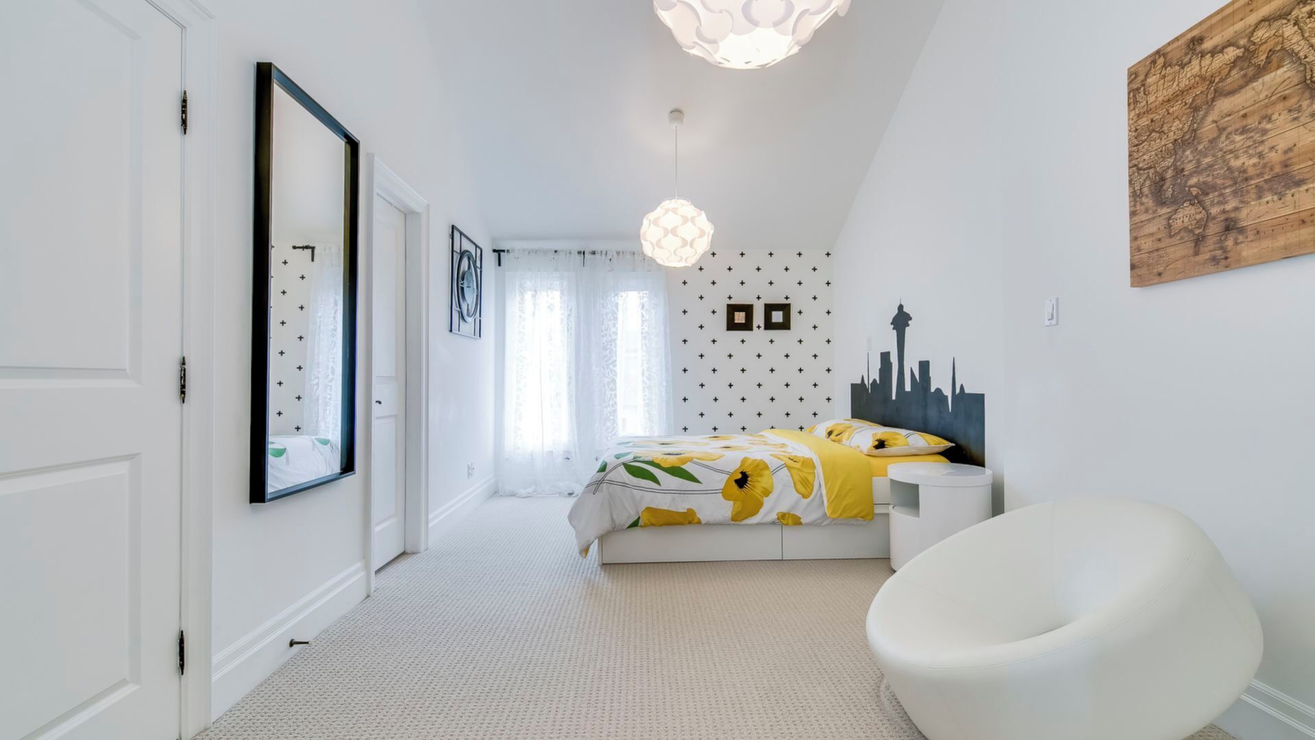 Une chambre avec un lit, une chaise blanche, un luminaire fixé au mur et une fresque représentant un paysage urbain.