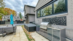 Cocina exterior con parrilla, zona de estar y vista a un barrio residencial.