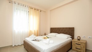 Una cama bien hecha con sábanas blancas, una mesita de noche de madera con cajones y una ventana con cortinas traslúcidas.