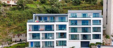 A modern multi-story building with balconies, a covered entrance, and a landscaped area with plants.