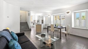 Una sala moderna con una mesa de centro de vidrio, un sofá con cojines, un área de comedor con sillas y una mesa, y una cocina al fondo.