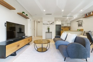 Una sala de estar moderna con televisor de pantalla plana, consola de madera, mesa de centro redonda, sofá gris y una zona de cocina al fondo.