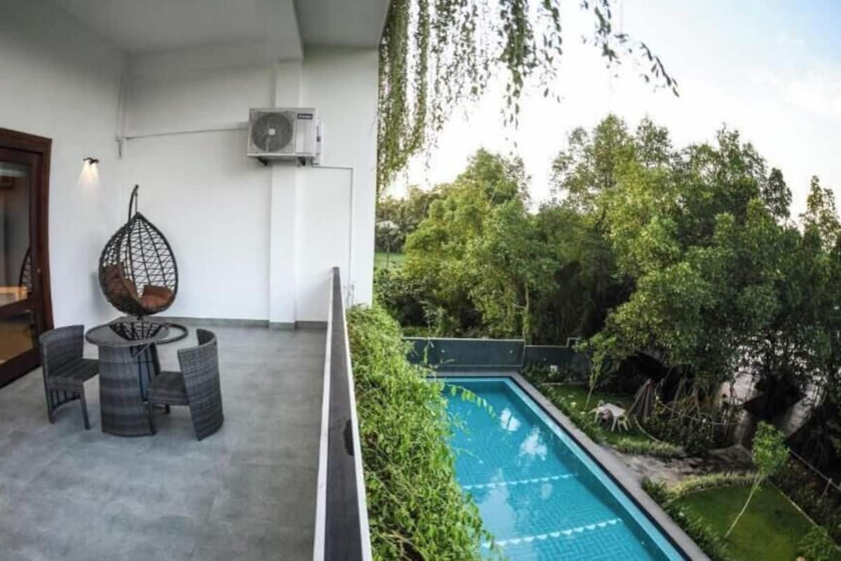 Un balcón con mesa y sillas, un equipo de aire acondicionado y vistas a una piscina rodeada de vegetación.