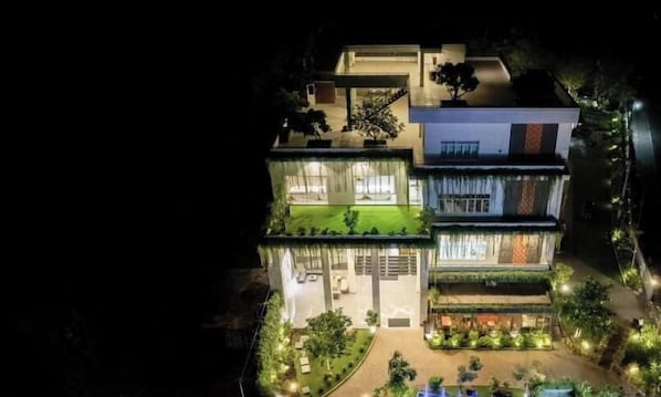 Un edificio moderno de varios pisos con una entrada bien iluminada, un área de estar exterior visible y un jardín en la azotea.