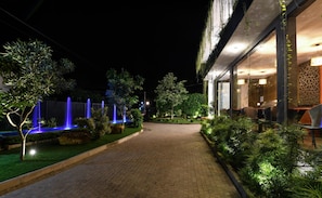 Un espacio al aire libre nocturno con un camino de ladrillo, plantas iluminadas y una fuente con luz azul.