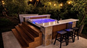 Ofurô ao ar livre com revestimento de pedra e madeira, área de bar com banquetas e luzes de fada em um jardim.