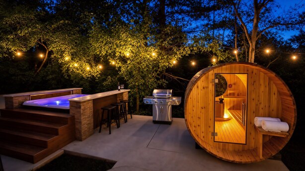 Eine holzverkleidete, fassförmige Sauna mit Sitzbank, ein Whirlpool mit blauem Licht und ein Grill im Freien.