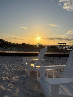 Zwei weiße Adirondack-Stühle an einem sandigen Strand mit einem Holzsteg und einem Pavillon im Hintergrund.