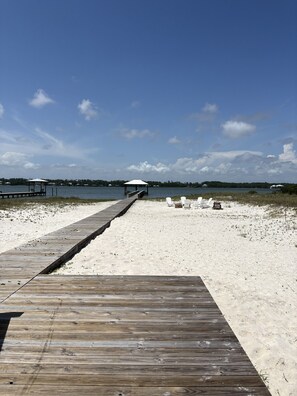 Eine Holzstege führt zu einem Strand mit einem Pavillon und Liegestühlen.