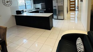 A modern kitchen with a central island, a dining area with a black and white sofa, and a hallway leading to a door. - Peaceful corner retreat|Park Side | 3 Bed, 2 Bath (Cranbourne West)