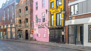 Eine Straße mit Gebäuden, darunter ein rosafarbenes Gebäude mit der Aufschrift 'Lucy Lounge' und ein gelbes Gebäude mit der Aufschrift 'Restaurant & Wine Bar'.