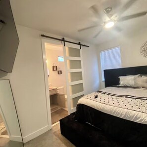 Ein Schlafzimmer mit Bett, Deckenventilator, Fenster mit Jalousien und einer Tür zu einem Badezimmer.