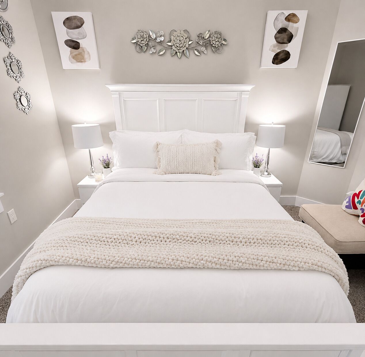 A white bedroom with a bed, two bedside tables, lamps, a decorative wall piece, and a mirror.