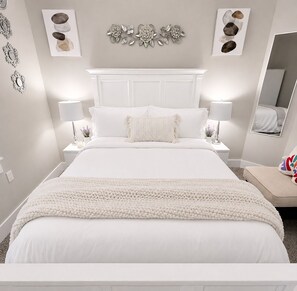 A white bedroom with a bed, two bedside tables, lamps, a decorative wall piece, and a mirror.