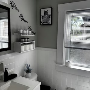 Casa de banho com sanita, lavatório e janela com persianas. Há prateleiras com artigos de higiene pessoal e um quadro com um gato.