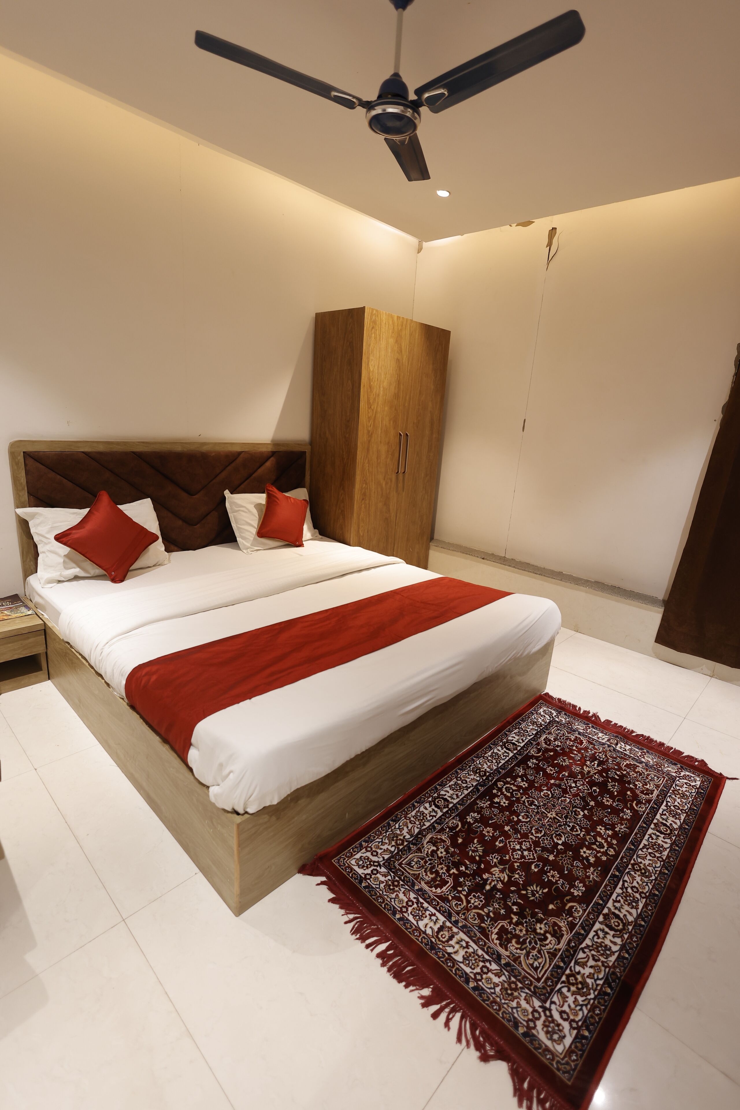 Une chambre à coucher avec un lit, une armoire en bois, un ventilateur de plafond et un tapis à motifs.