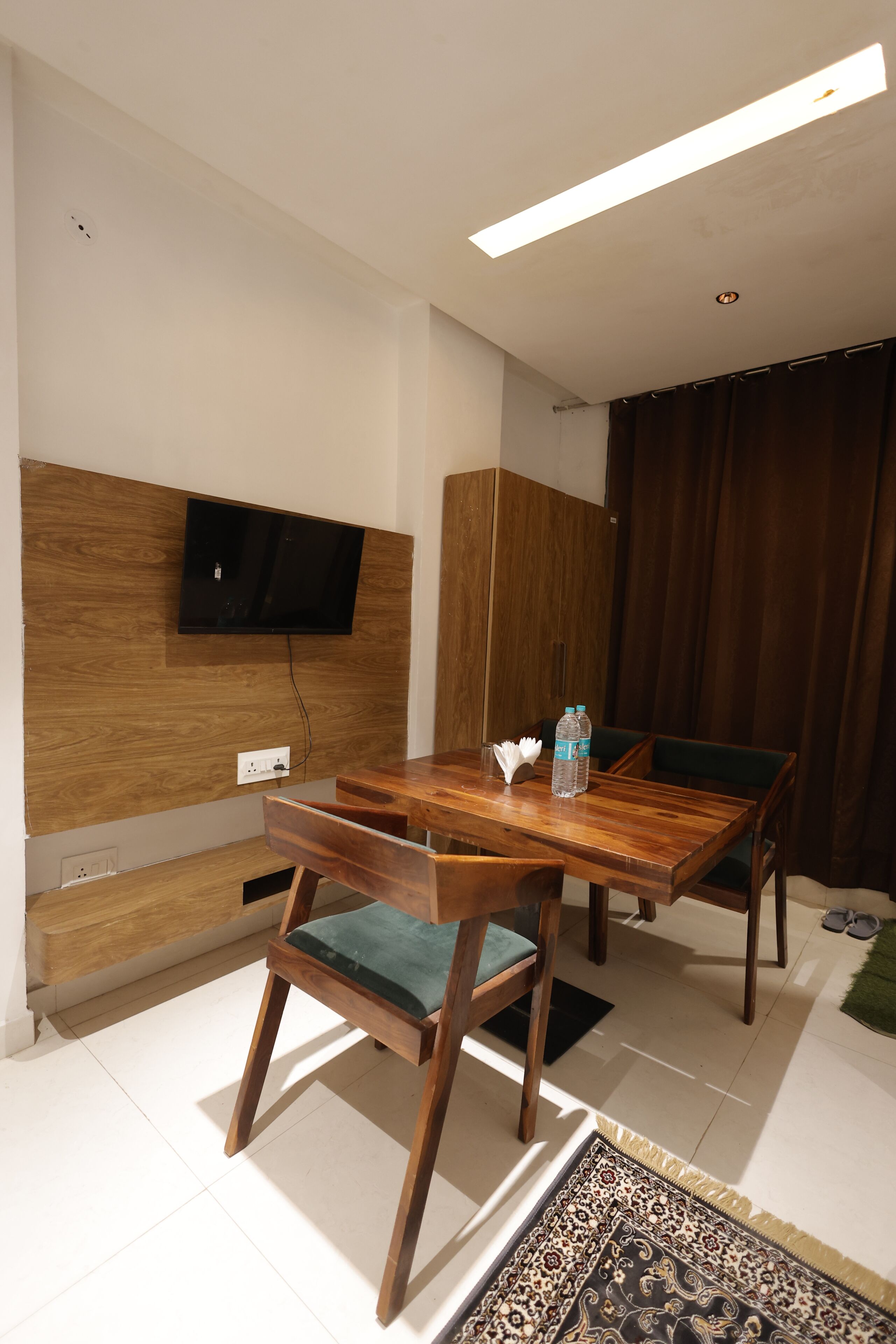 A modern room with a wooden dining table, two chairs, a flat-screen TV mounted on a wooden panel, a wardrobe, and a large window with curtains.
