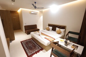 A hotel room with a bed, a desk, a chair, a sofa, a ceiling fan, and an air conditioning unit.
