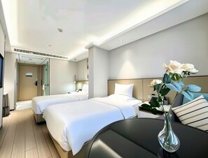 Habitación de hotel con dos camas, un escritorio, una silla, un florero con flores y una puerta que da a un baño.