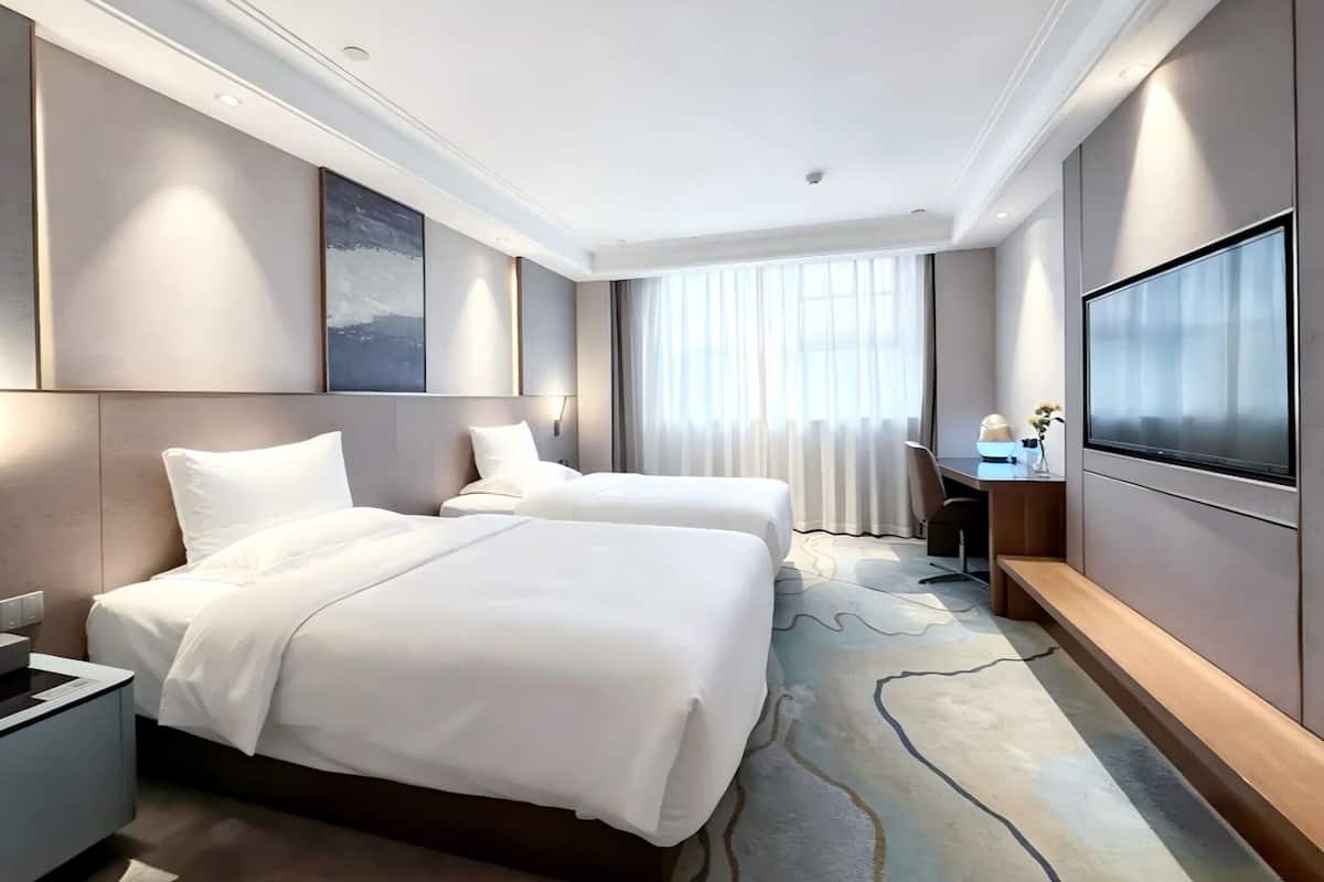 Ein Hotelzimmer mit zwei Betten, einem Schreibtisch mit Stuhl, einem Flachbildfernseher und einem großen Fenster mit Vorhängen.