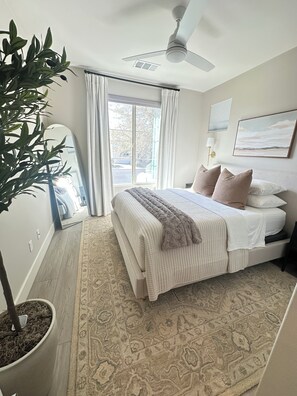 Ein Schlafzimmer mit einem Bett, einem Deckenventilator, einem Fenster mit Vorhängen, einer Zimmerpflanze und einem Teppich.