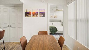 A dining area with a wooden table and chairs, a white wall with three paintings, a white door, and a bed with a green blanket in the background. - Central Scottsdale Haven Condo (Scottsdale)