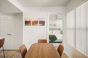 Ein Essbereich mit Holztisch und -stühlen, einer weißen Wand mit drei Bildern, einer weißen Tür und einem Bett mit grüner Decke im Hintergrund.