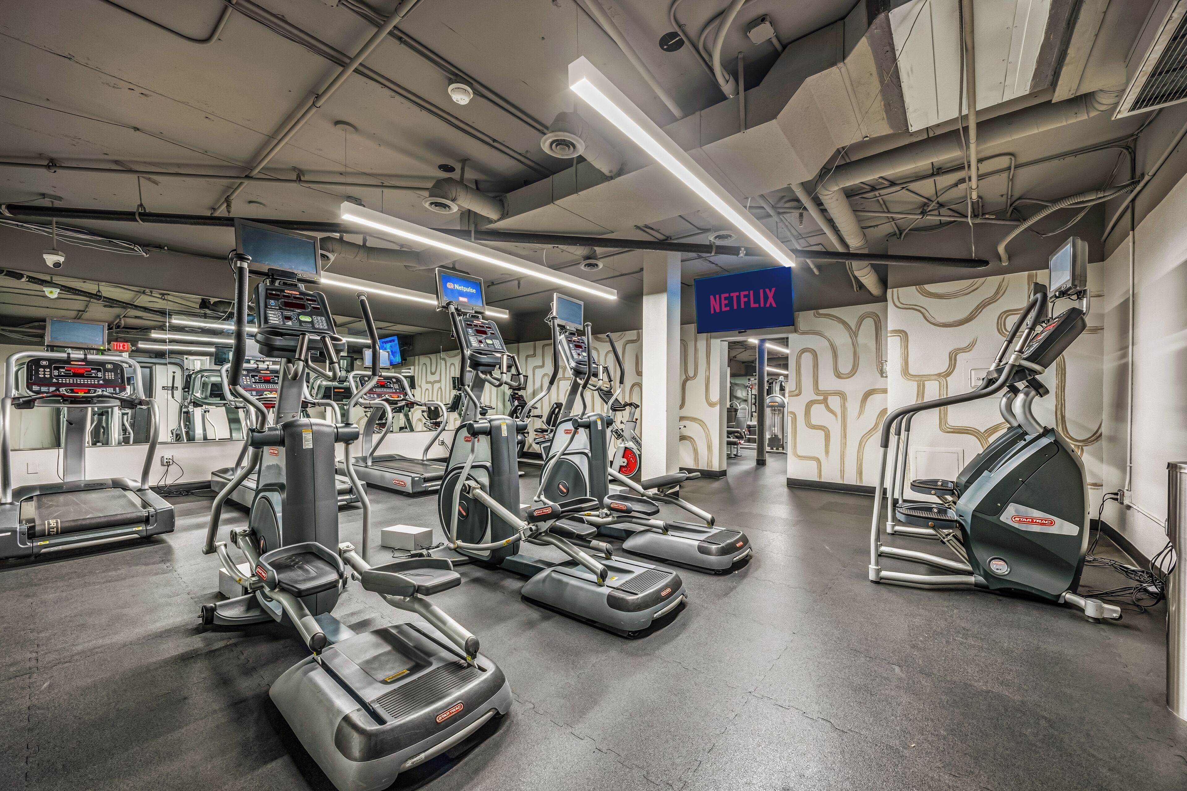 A gym with various cardio machines, a treadmill, and a wall-mounted TV screen.