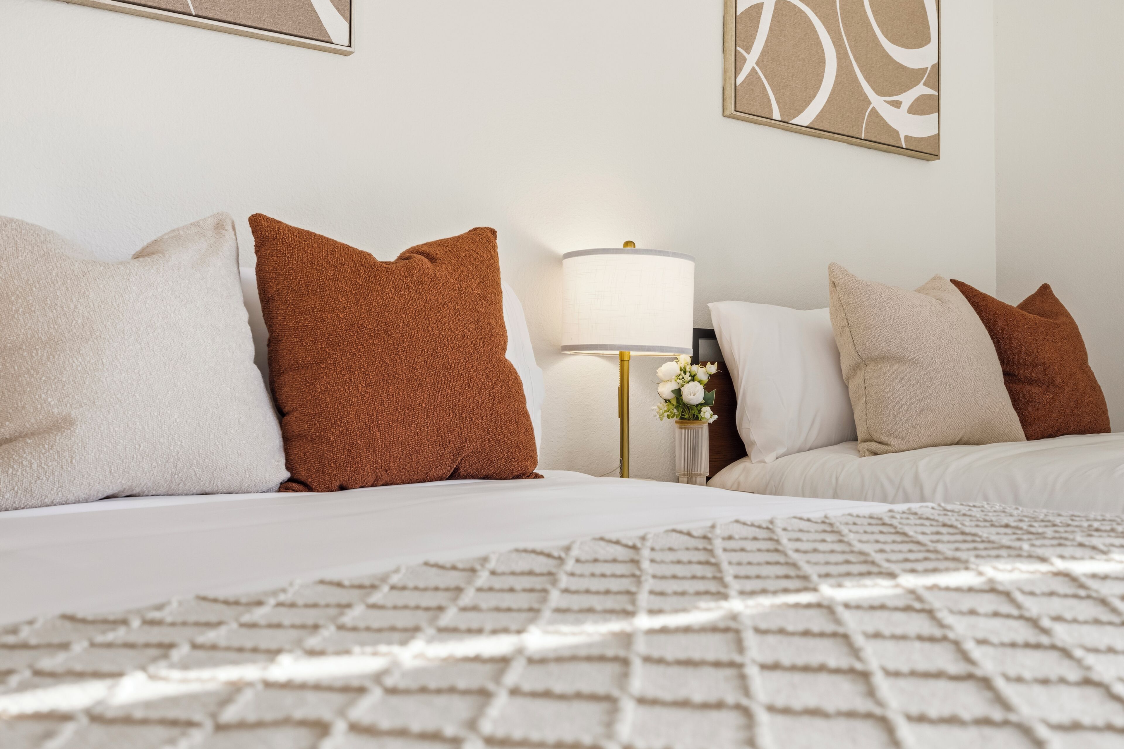 A neatly made bed with white and brown pillows, a white blanket, and a bedside table with a lamp and a vase of flowers.
