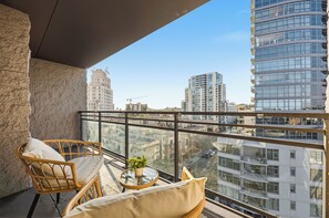 Uma varanda com móveis de vime, uma pequena mesa com uma planta e vista para prédios da cidade.