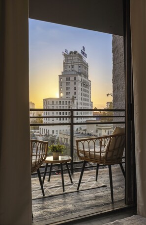 Ein Balkon mit zwei Korbstühlen und einem kleinen Tisch, mit Blick auf eine Stadtlandschaft, in der ein markantes Gebäude mit der Aufschrift 'CONNER' zu sehen ist.