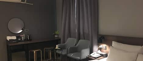 Habitación de hotel con una cama, un escritorio con silla, una mesita, un espejo y una ventana con cortinas.