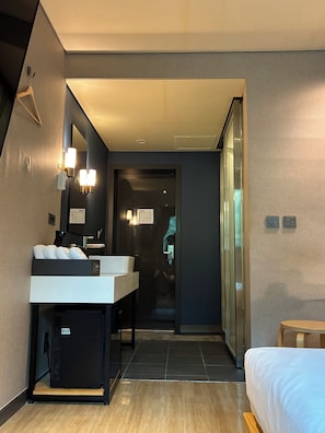A modern hotel room with a large bed, a bedside table, a built-in desk with a chair, a large mirror, and a wall-mounted light fixture.