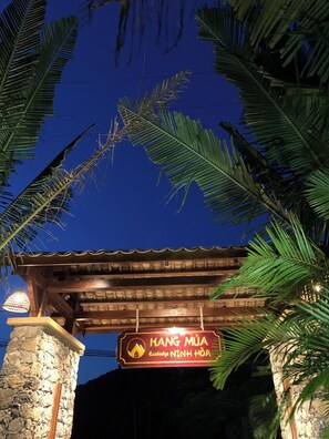 Señal para 'HANG MUÀ Ecolodge NINH HOA' con un pilar de piedra y hojas de palma enmarcando la entrada.