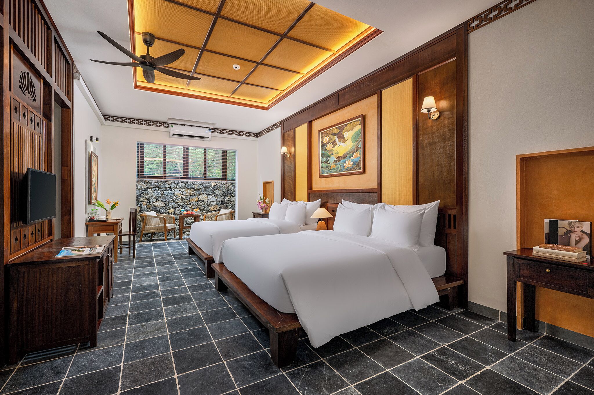 A hotel room with two beds, a ceiling fan, a stone wall, and a television.