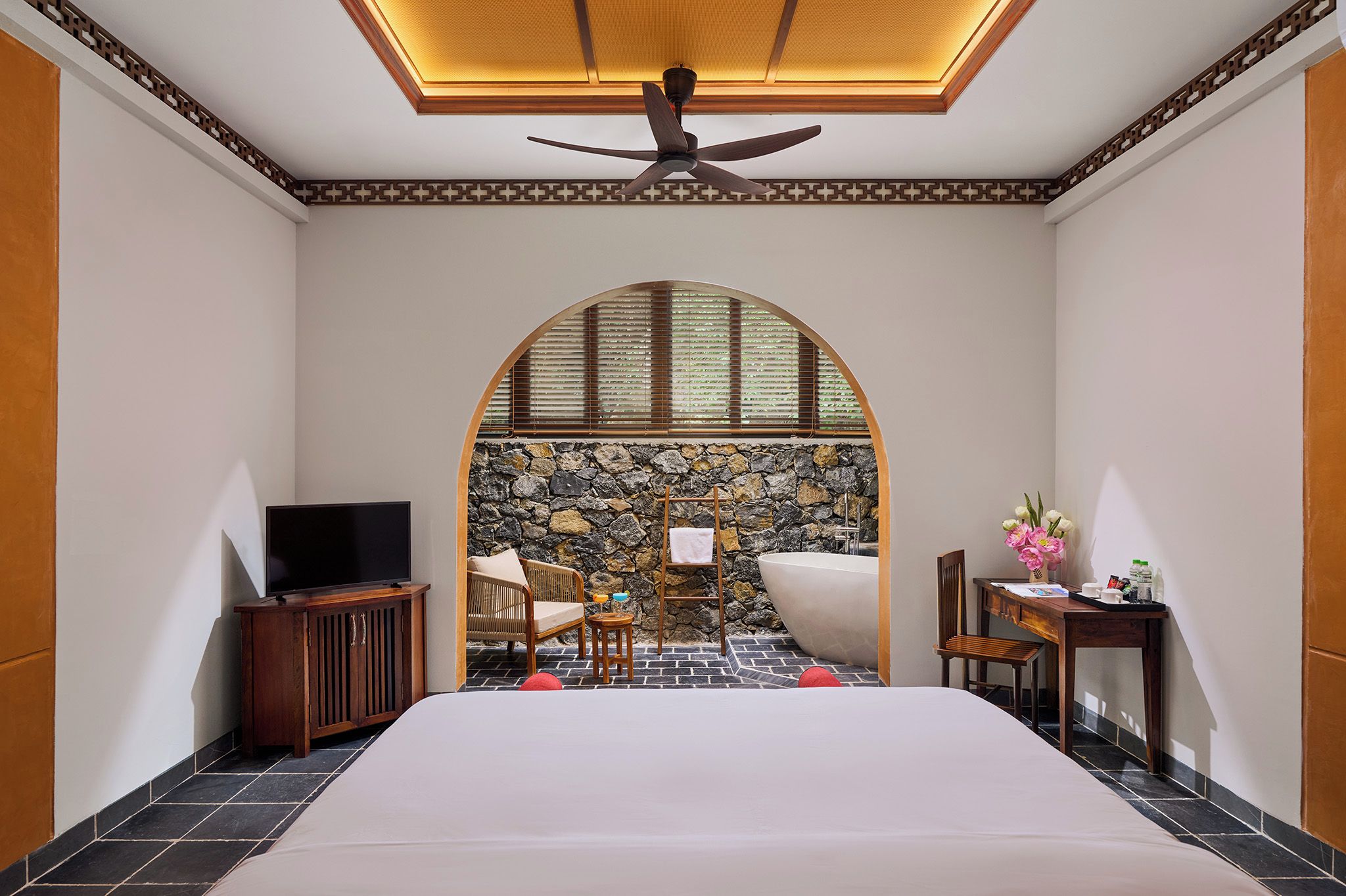 A hotel room with a large bed, a television, a seating area with a stone wall backdrop, and a ceiling fan.