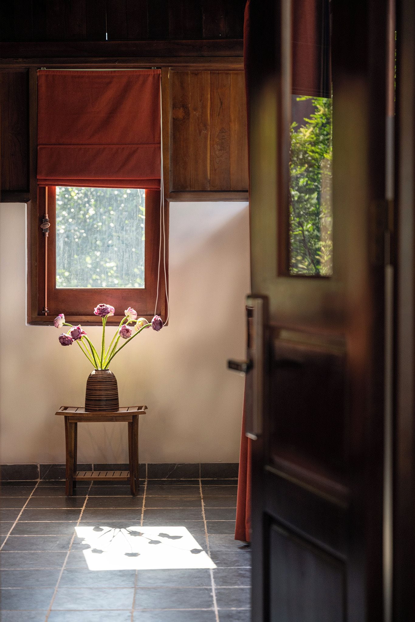 A room with a wooden door, a window with a view of greenery, a small table with a vase of flowers, and a tiled floor.