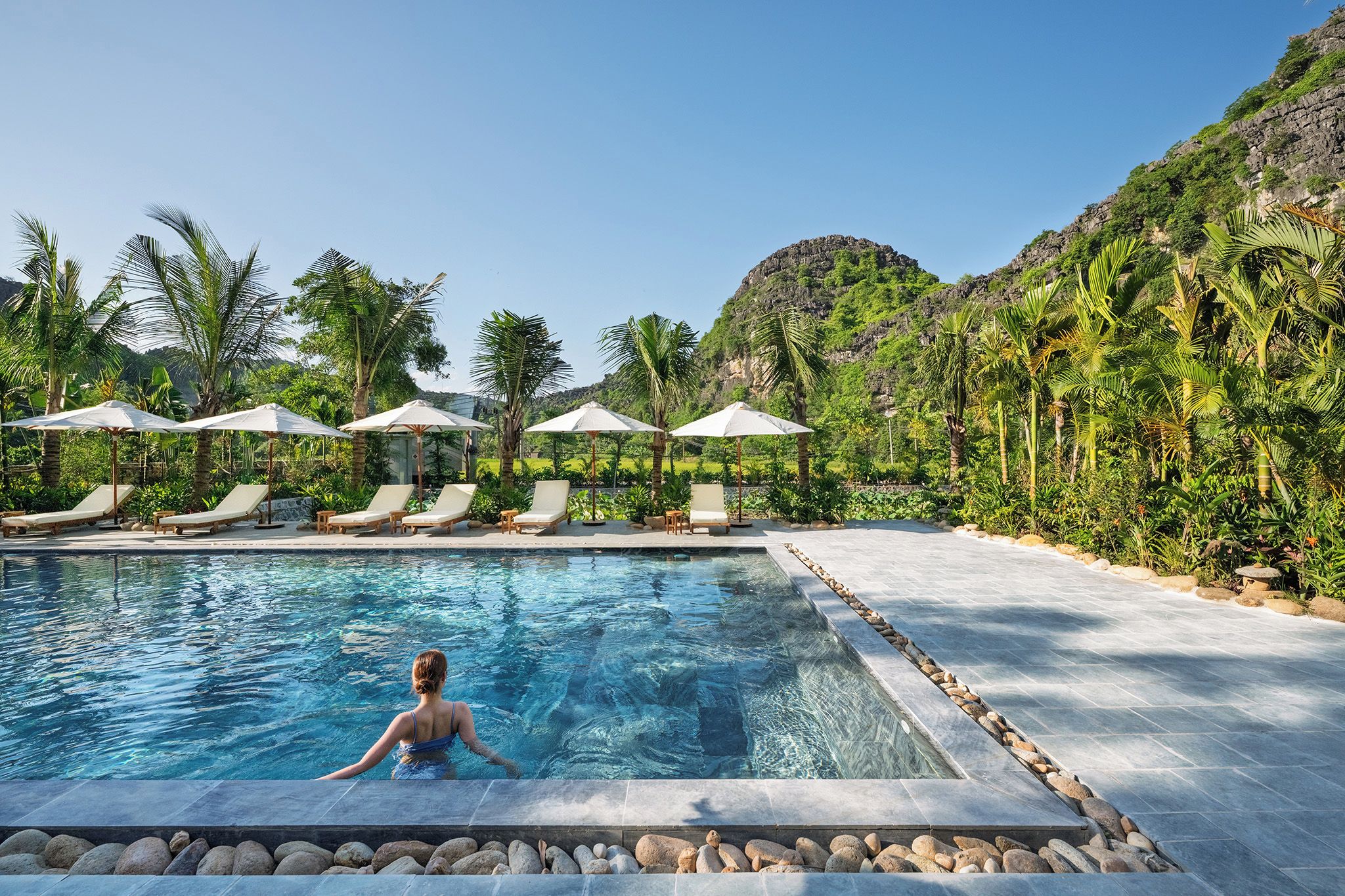 A swimming pool with lounge chairs and umbrellas, surrounded by lush greenery and a mountainous backdrop.