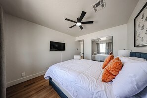 Ein Schlafzimmer mit einem Bett, einem Deckenventilator, einem Fernseher, einem Spiegel und einer Tür zu einem anderen Raum.