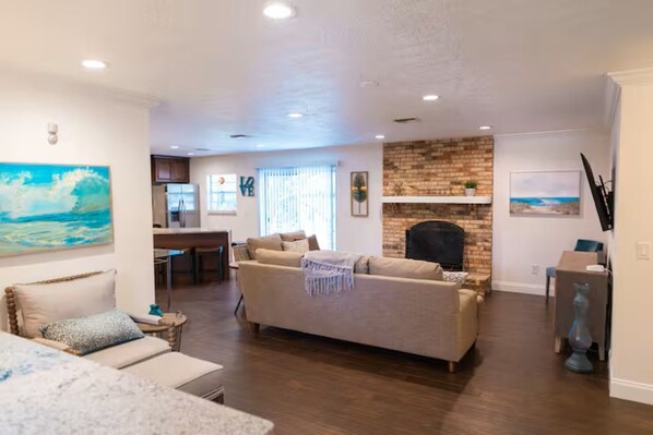 A living room with a fireplace, a sofa, a dining area, and a kitchen in the background. - Harbor Home (Oldsmar)