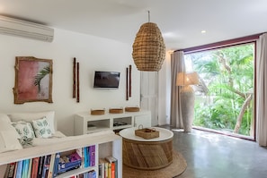 A modern living room with a sofa, bookshelves, a TV, a woven light fixture, and a view of greenery through a large window.
