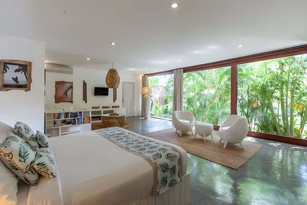 A modern bedroom with a large bed, a TV, a bookshelf, and a view of lush greenery. -  Kima Surf Camp Bali Canggu (Canggu)